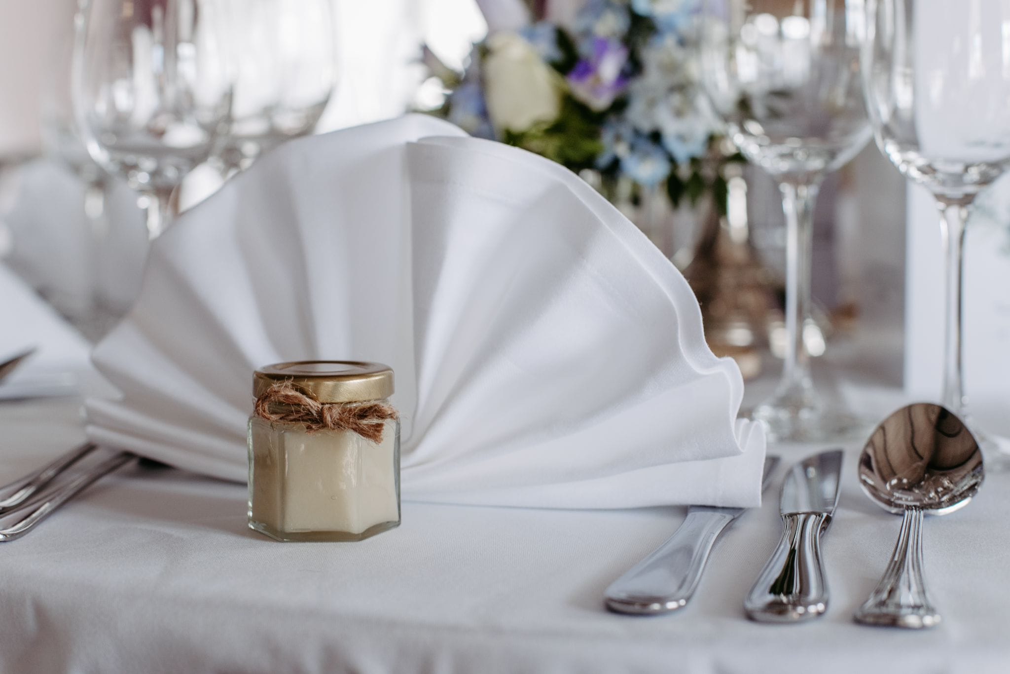 Ein Gastgeschenk in einem kleinen Honigglas mit goldenem Deckel und eine Serviette auf einem festlich gedeckten Tisch.